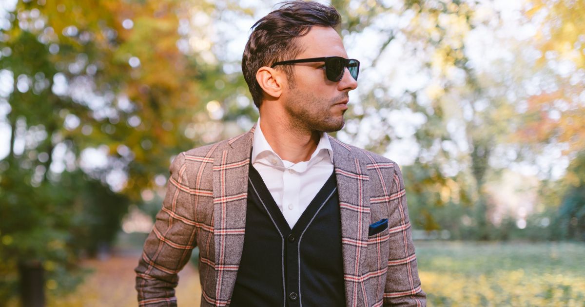 Man in plaid blazer and sunglasses in park with autumn foliage.
