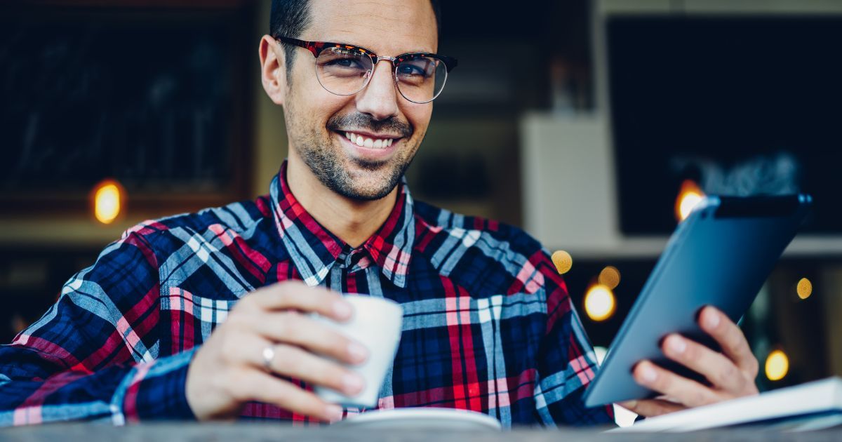 A man wearing glasses is sitting at a table holding a cup of coffee and a tablet.