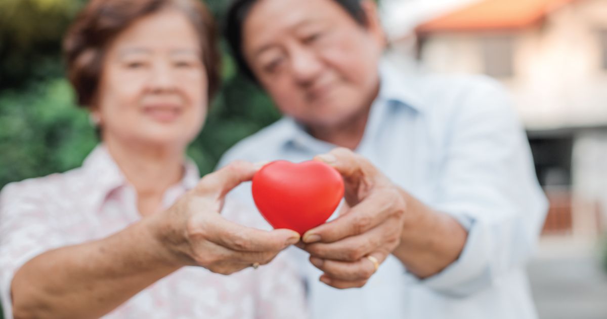 Hands holding a small red heart-shaped object in the foreground, with a smiling couple blurred behind.