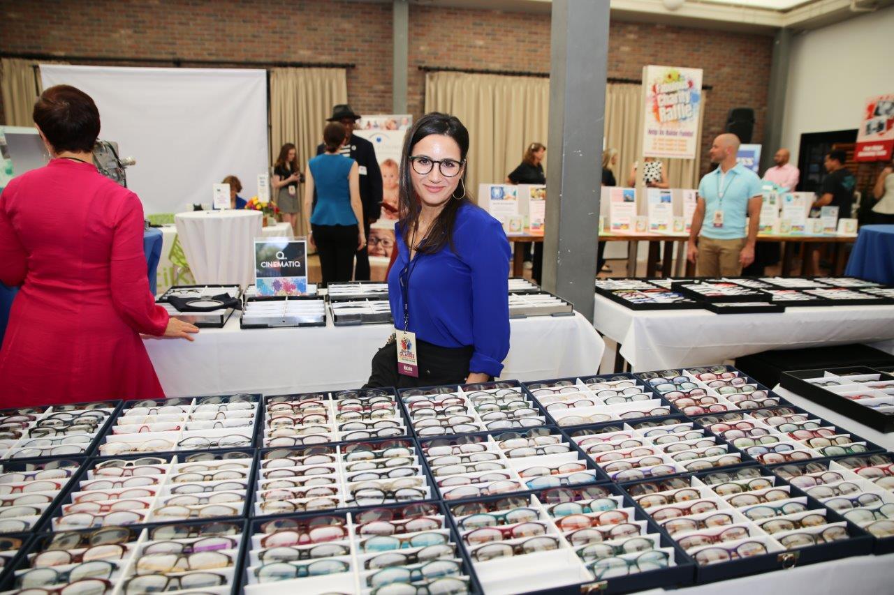 Vendor wearing glasses and standing behind tabletop eyewear display at 2018 Raise Your Glasses