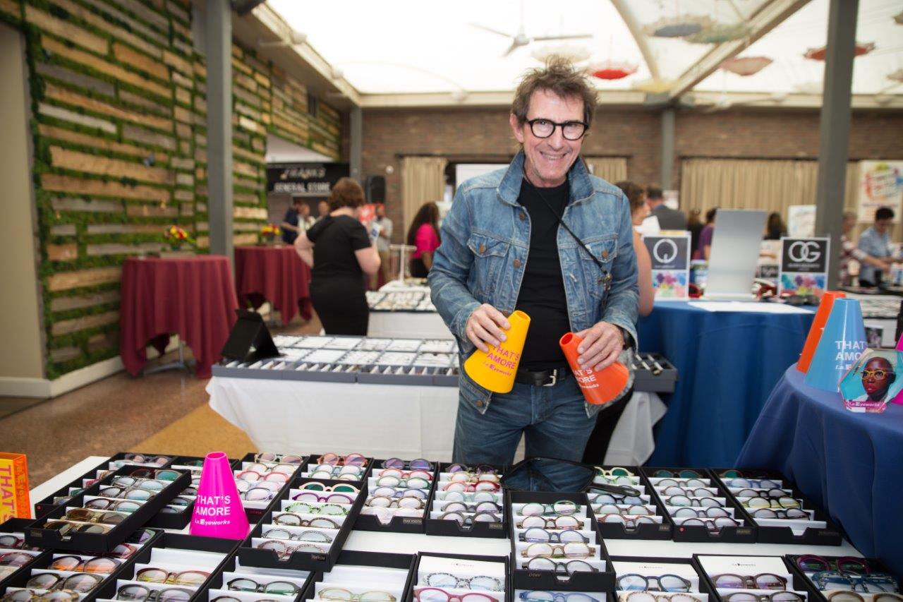 Smiling vendor wearing glasses and standing behind tabletop display at 2018 Raise Your Glasses