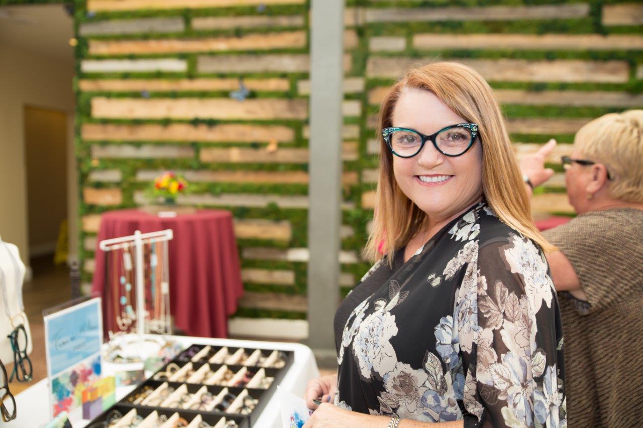 Smiling person wearing glasses and standing in front of tabletop display at 2018 Raise Your Glasses