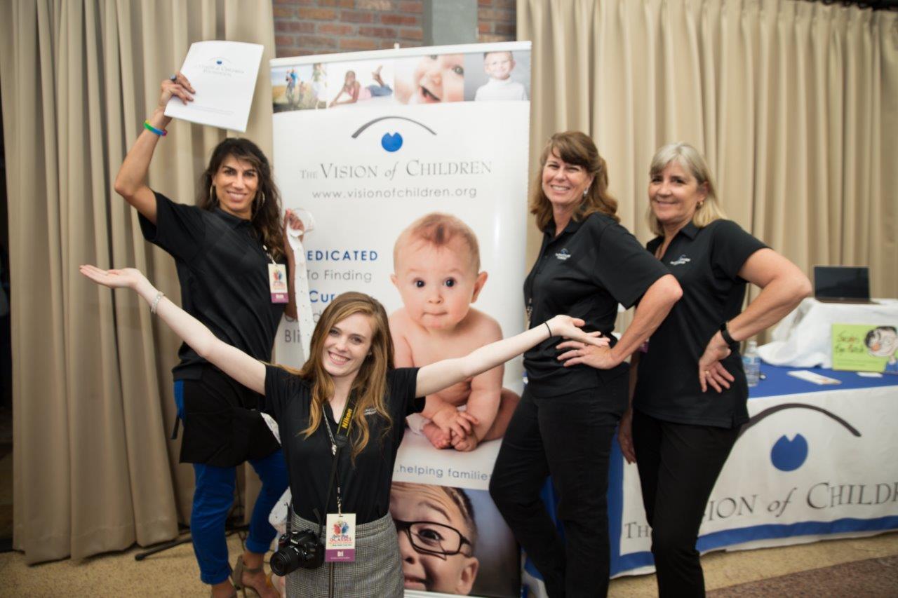 4 people standing in front of large banner at Raise Your Glasses 2018 Event