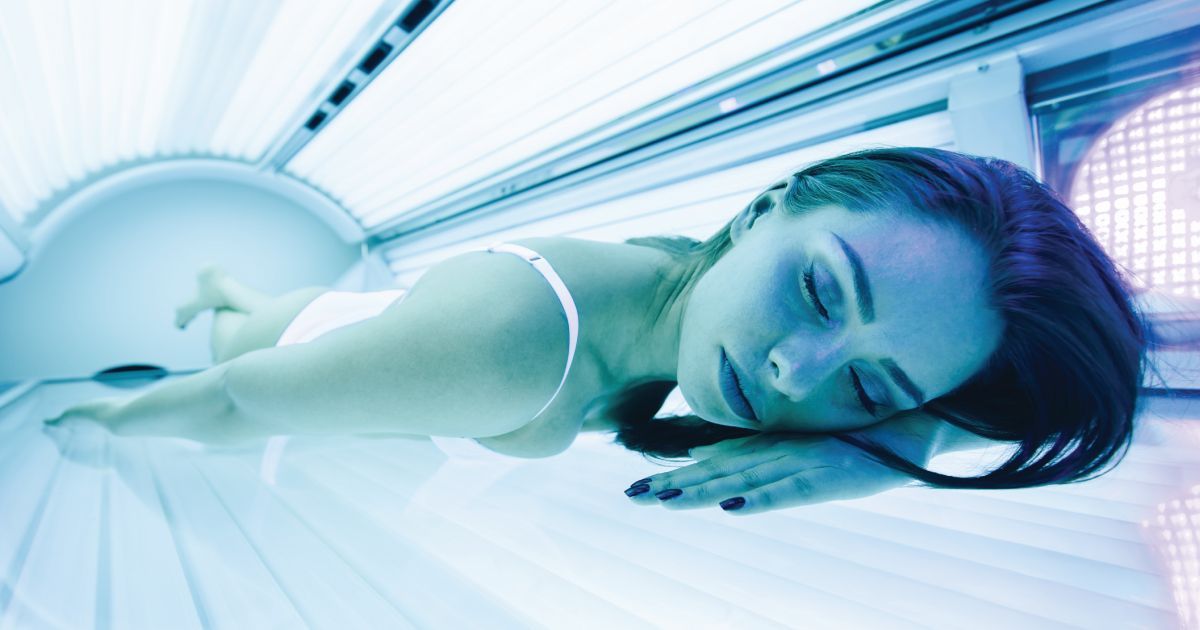 Woman lying inside an indoor tanning bed illuminated by blue UV light with eyes closed during a tanning session