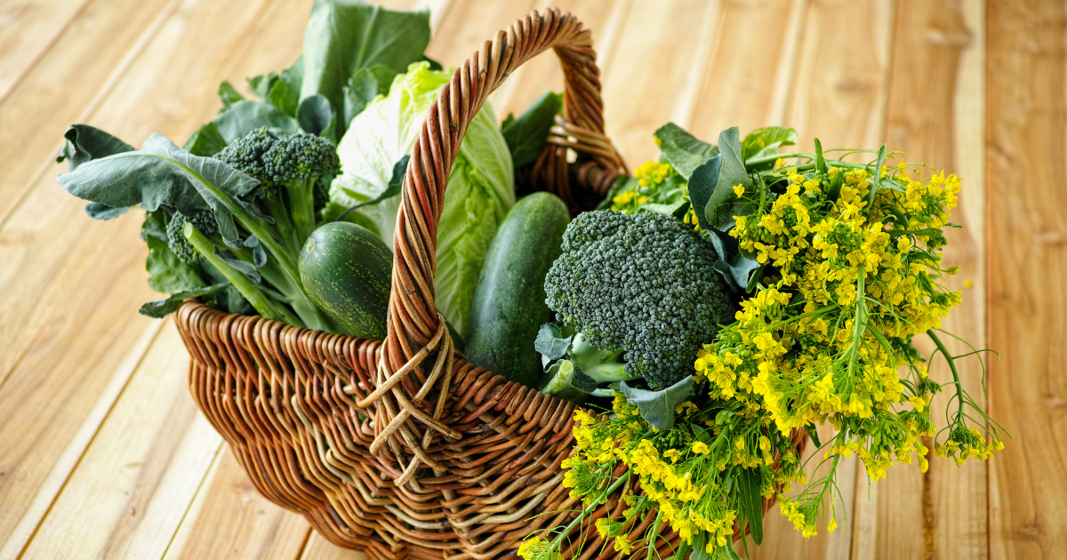 A wicker basket filled with green vegetables and yellow flowers on a wooden table