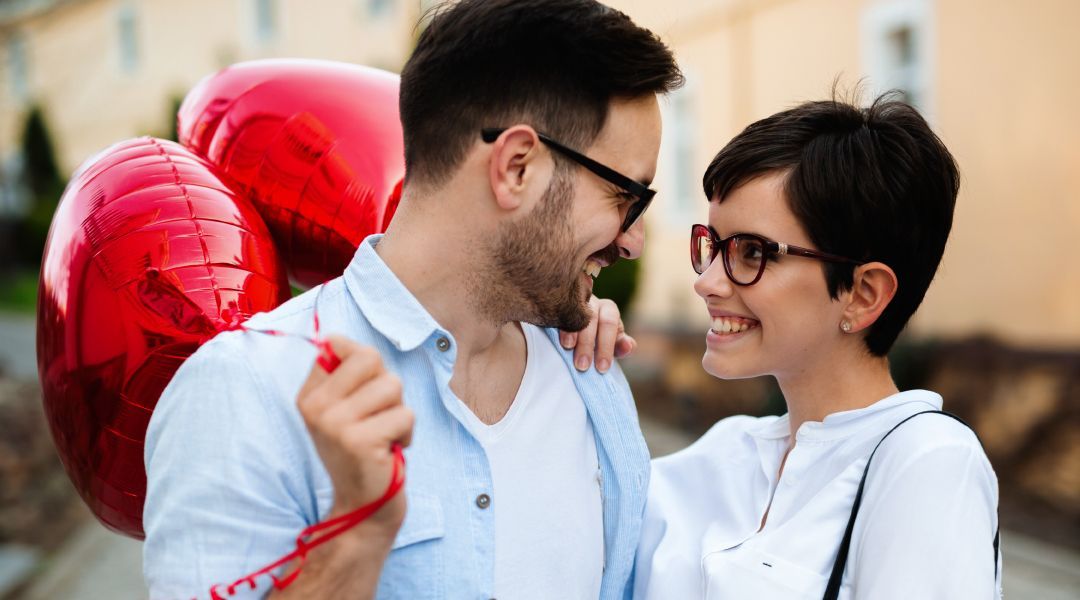 A man and a woman are looking at each other while holding red heart shaped balloons