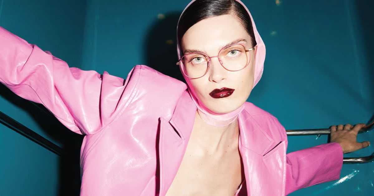 Person in a pink jacket and glasses posing against a teal background, arms outstretched.