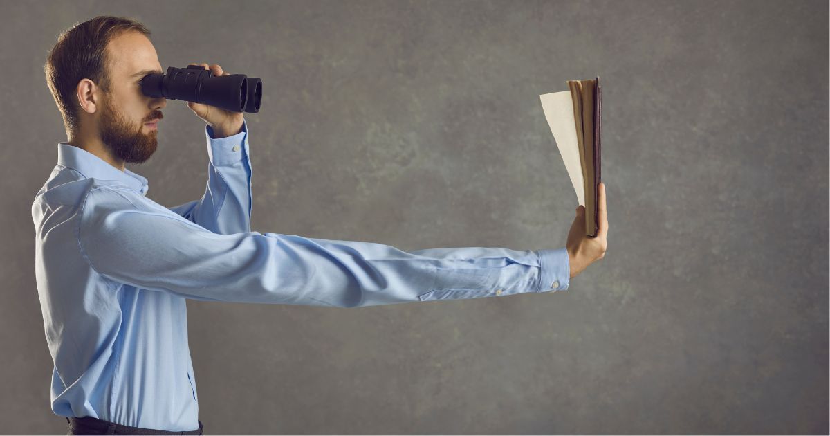 A man looking through binoculars while holding a piece of paper in his out-stretched arm.