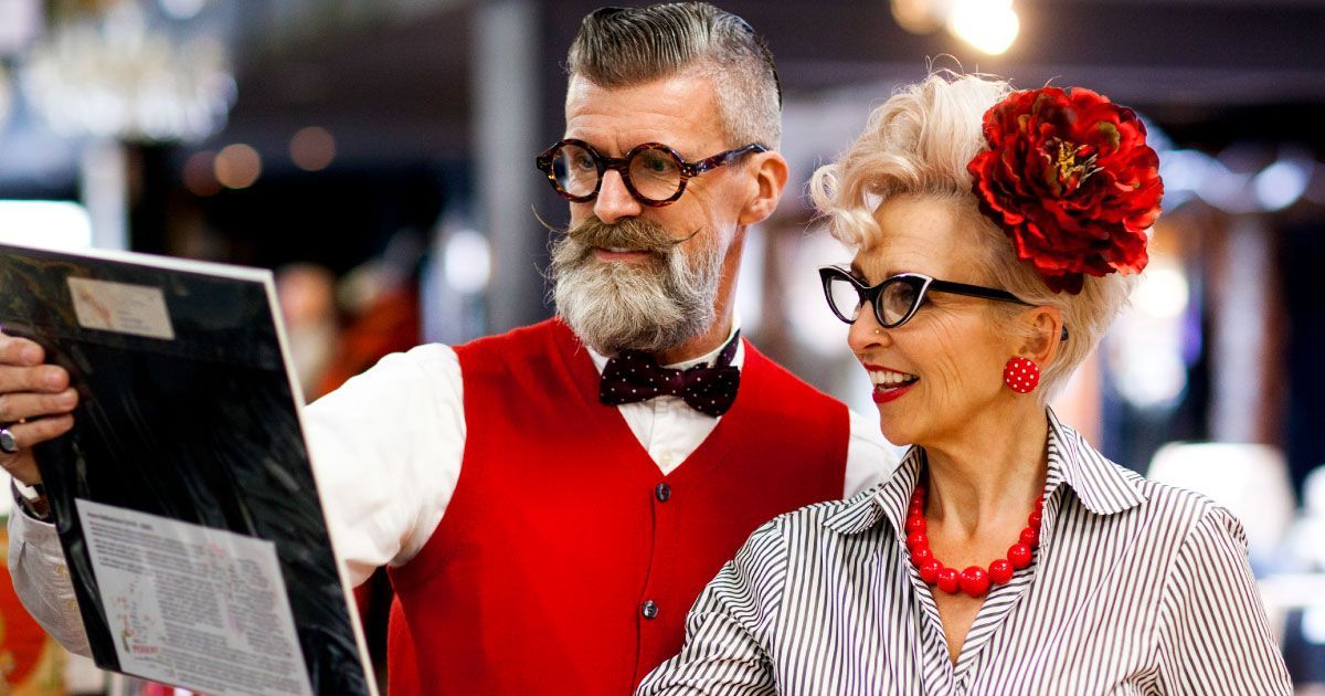 A man and a woman in retor clothing and glasses are looking at a menu in a restaurant.
