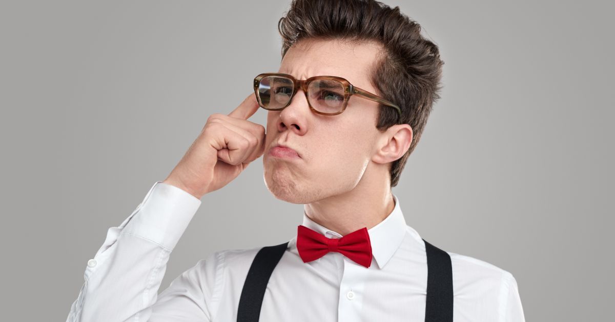 A young man wearing glasses and red bow tie has his finger to his temple and a questioning look.