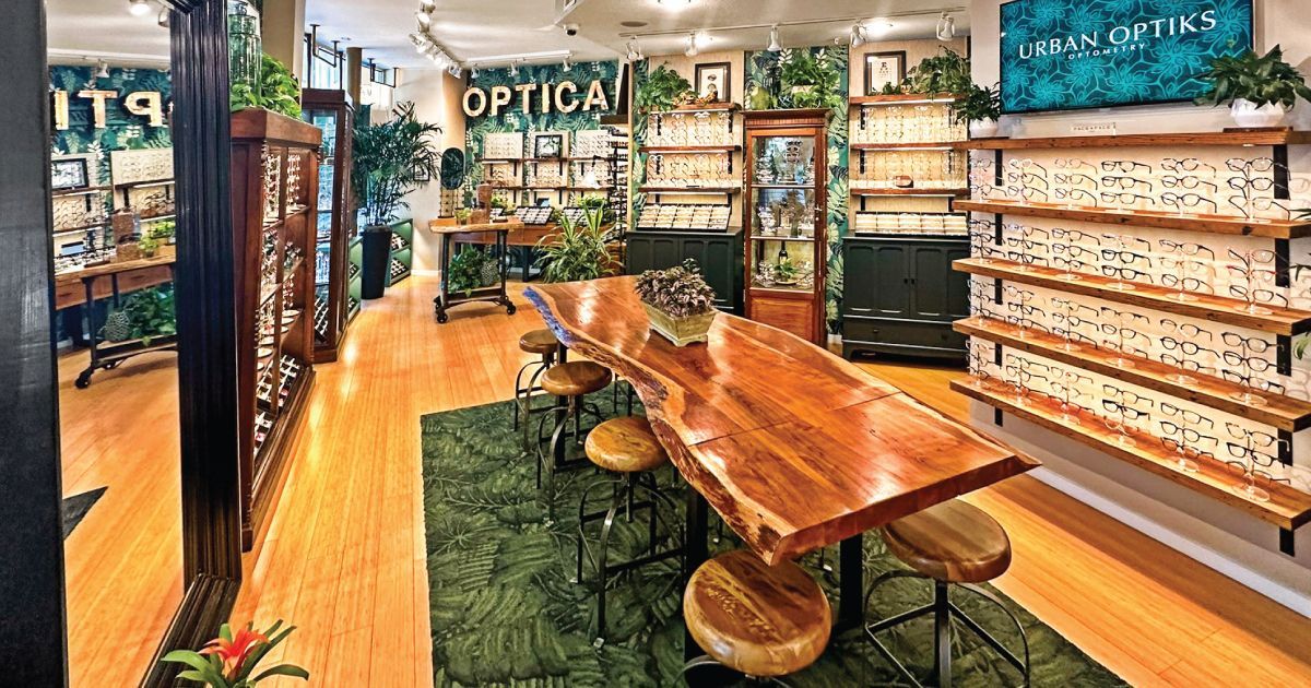 Interior phot of Urban Optiks Optometry with wooden table, green rug, shelves of eyeglass frames, and mirror displays.