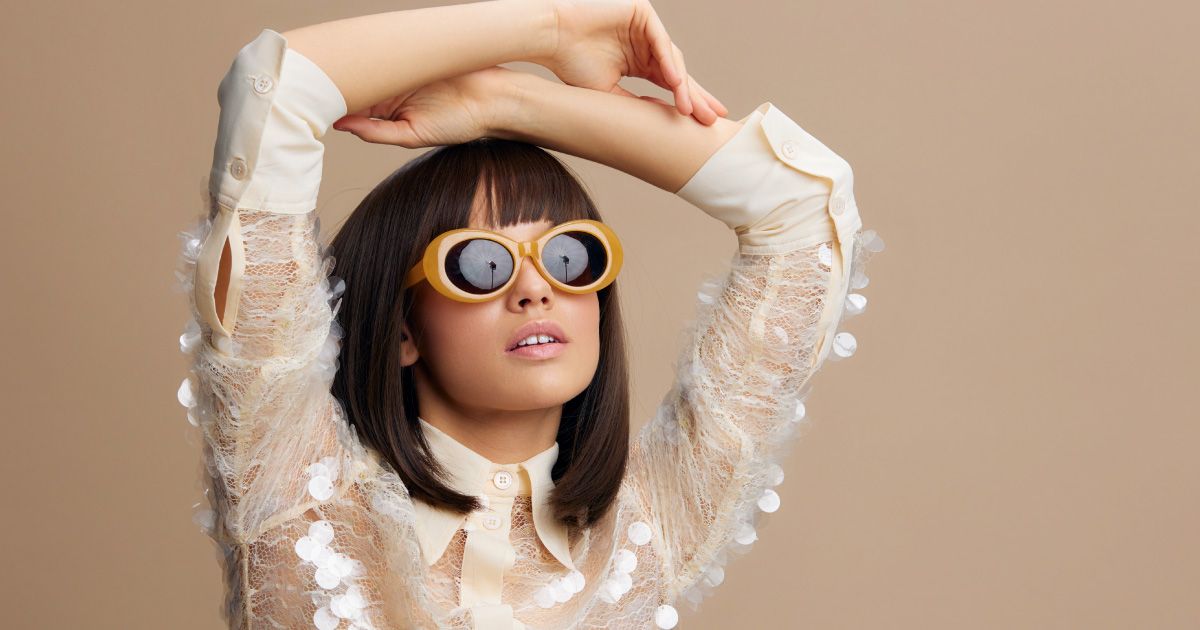 Woman wearing yellow-rimmed sunglasses and a cream-colored top with arms above her head.