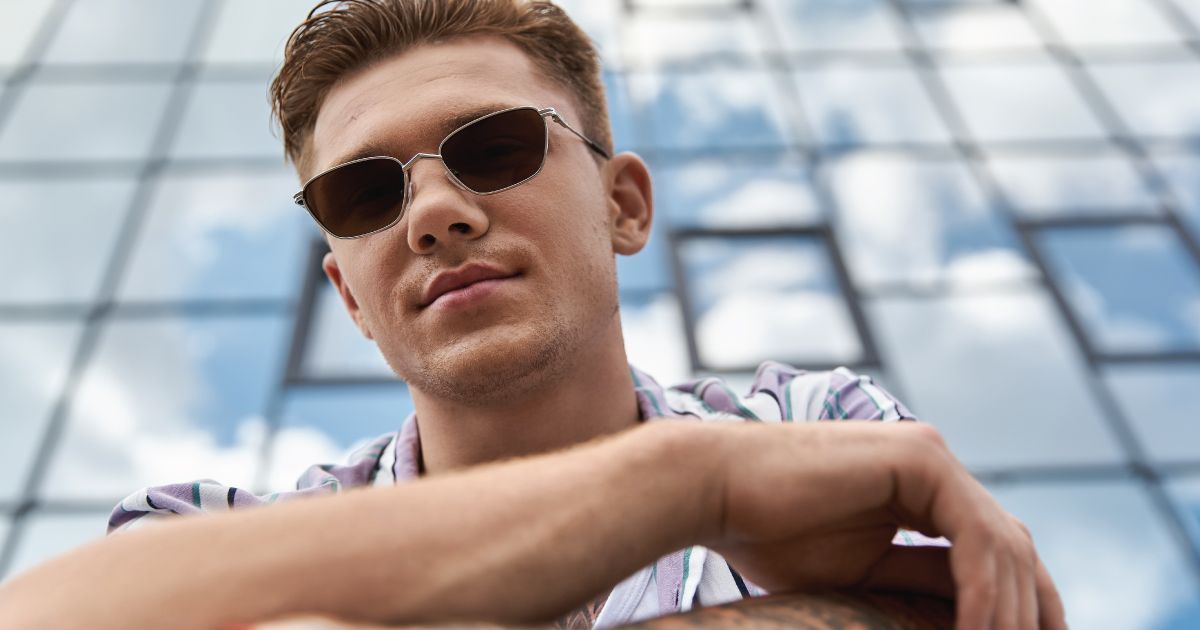 Young man wearing sunglasses, leaning on a railing in front of a glass building.