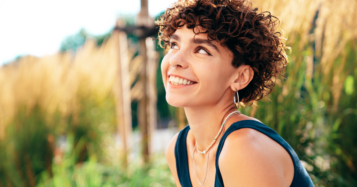 Smiling person with curly hair in a blue top looking up outdoors near greenery