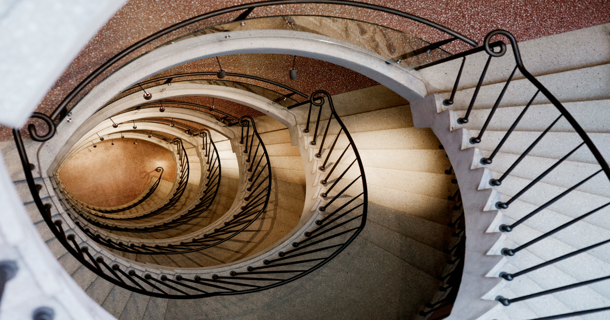 Spiral staircase viewed from above, with beige steps and black wrought-iron railings.