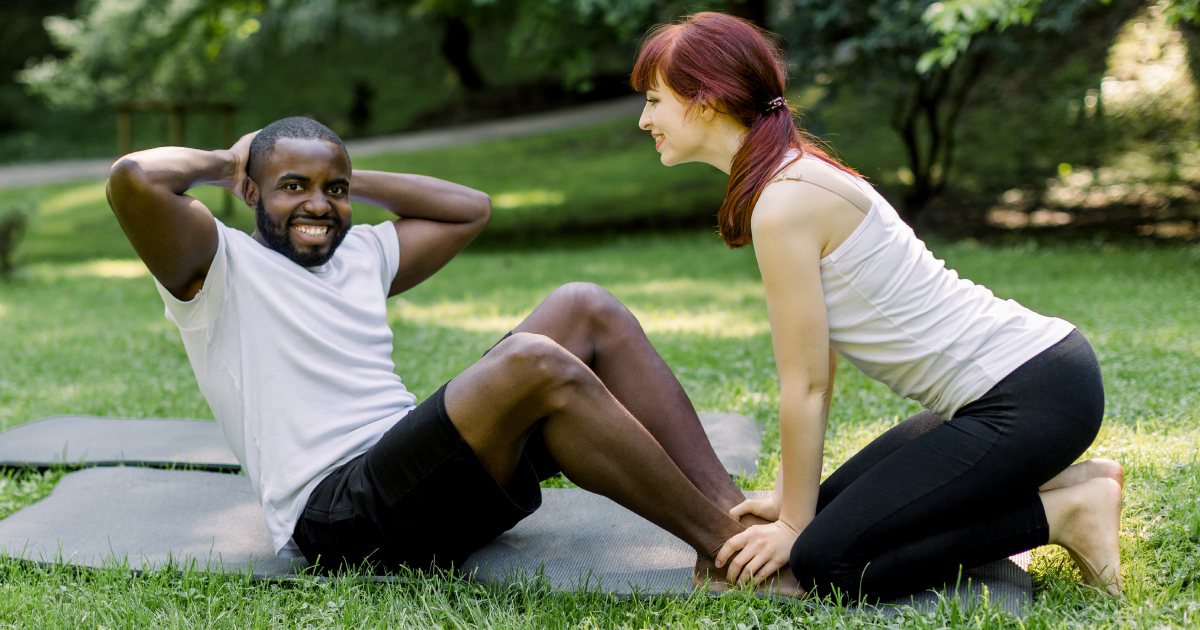 Two people doing sit-ups on exercise mats in a grassy park, one smiling and the other holding a pose.