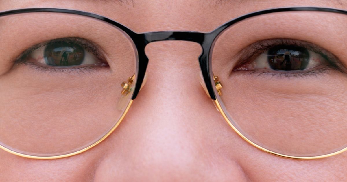 Close-up of a person's face wearing black and gold-rimmed glasses. Reflections in lenses, warm skin tone.