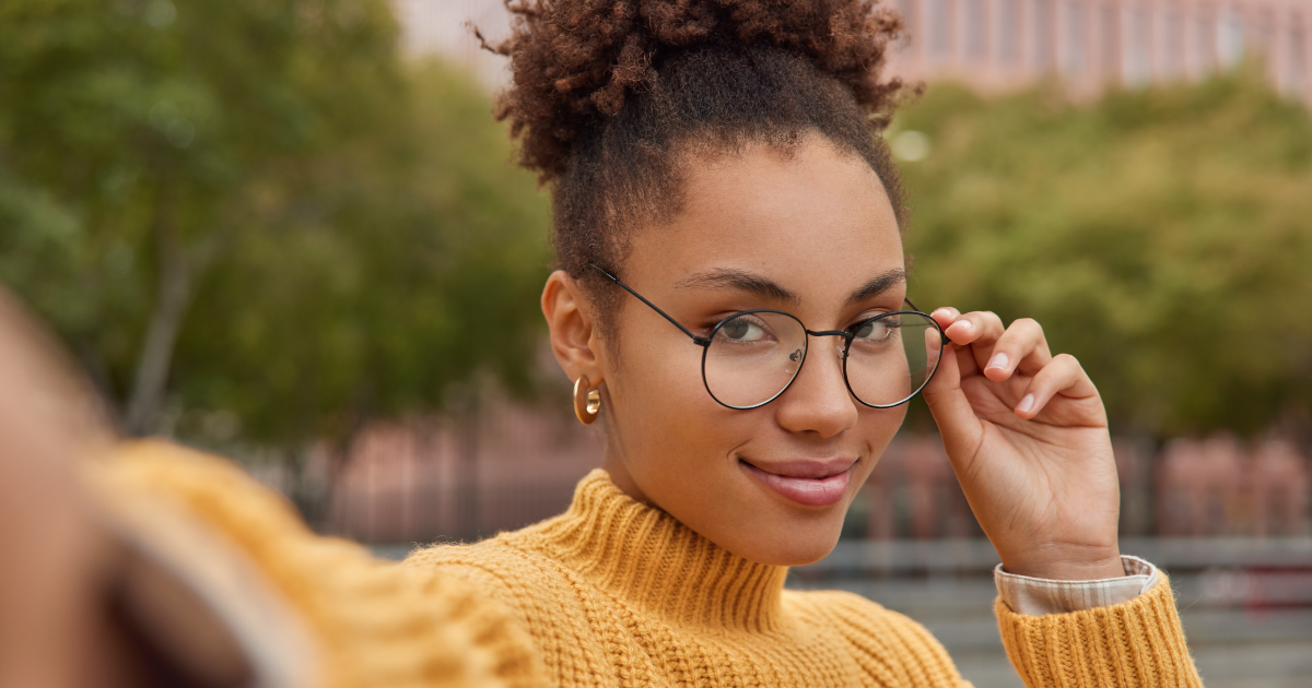Person wearing round glasses and a mustard sweater smiling outdoors, adjusting their glasses.