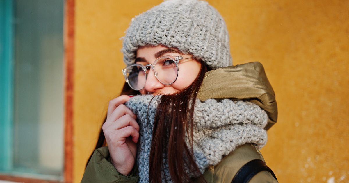 Woman in knit hat, scarf, and glasses, bundled against the cold, near a yellow wall.