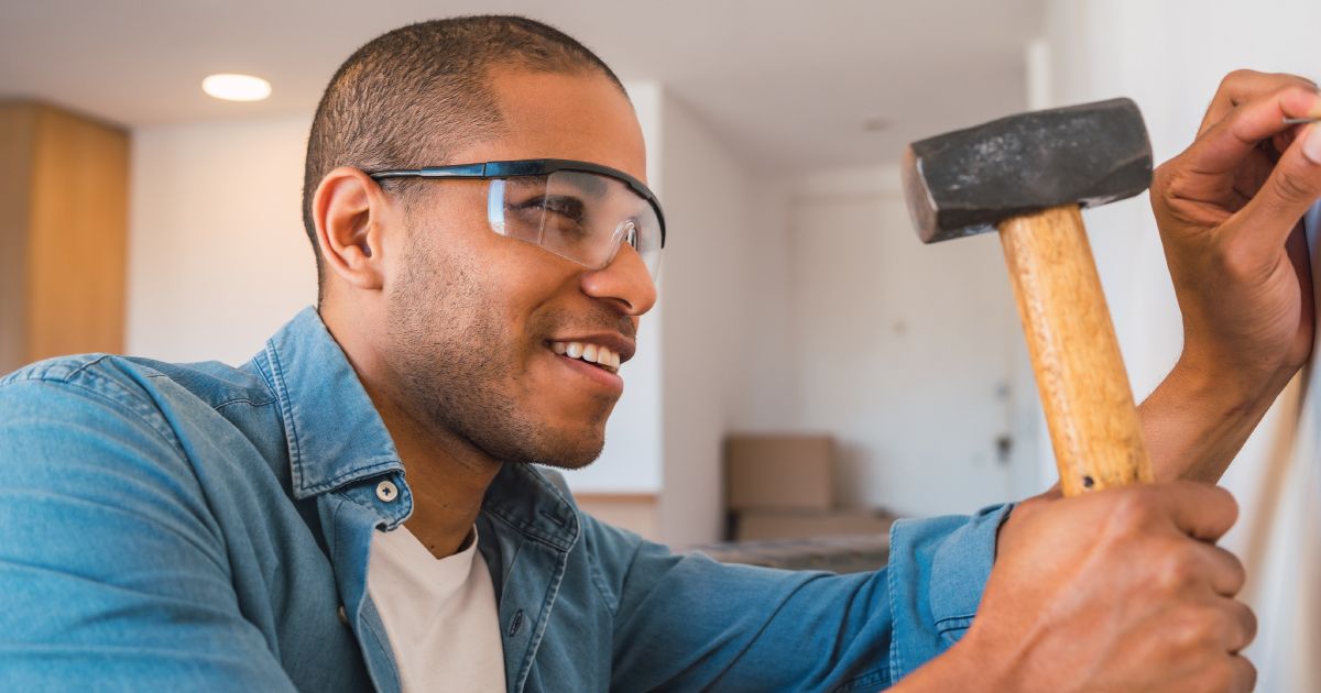 Man in safety glasses holding a hammer and nailing a nail into a wall
