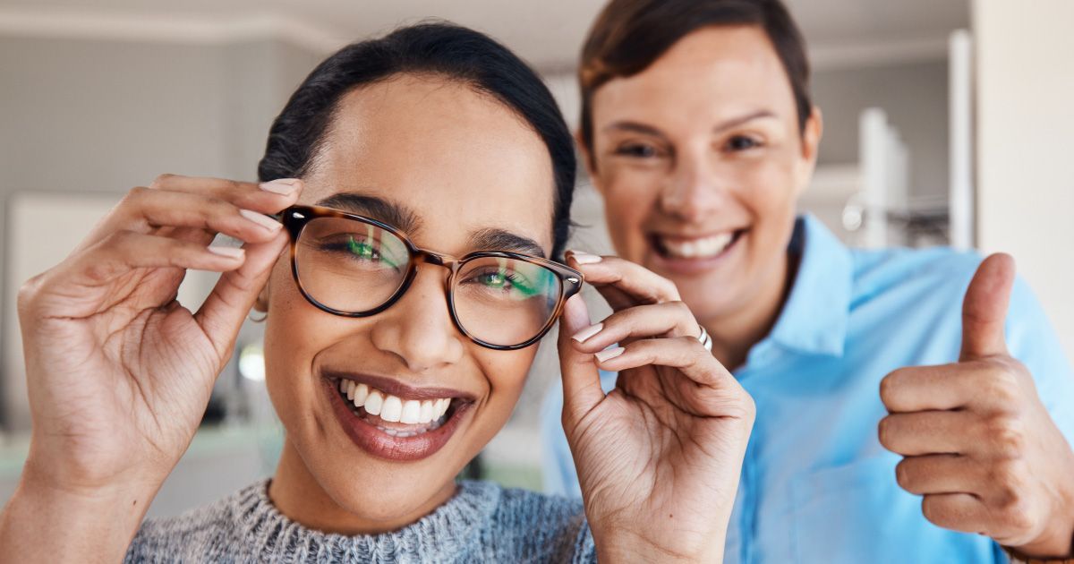 Woman wearing new glasses, smiling, with the optometrist standing behind her giving a thumbs up.