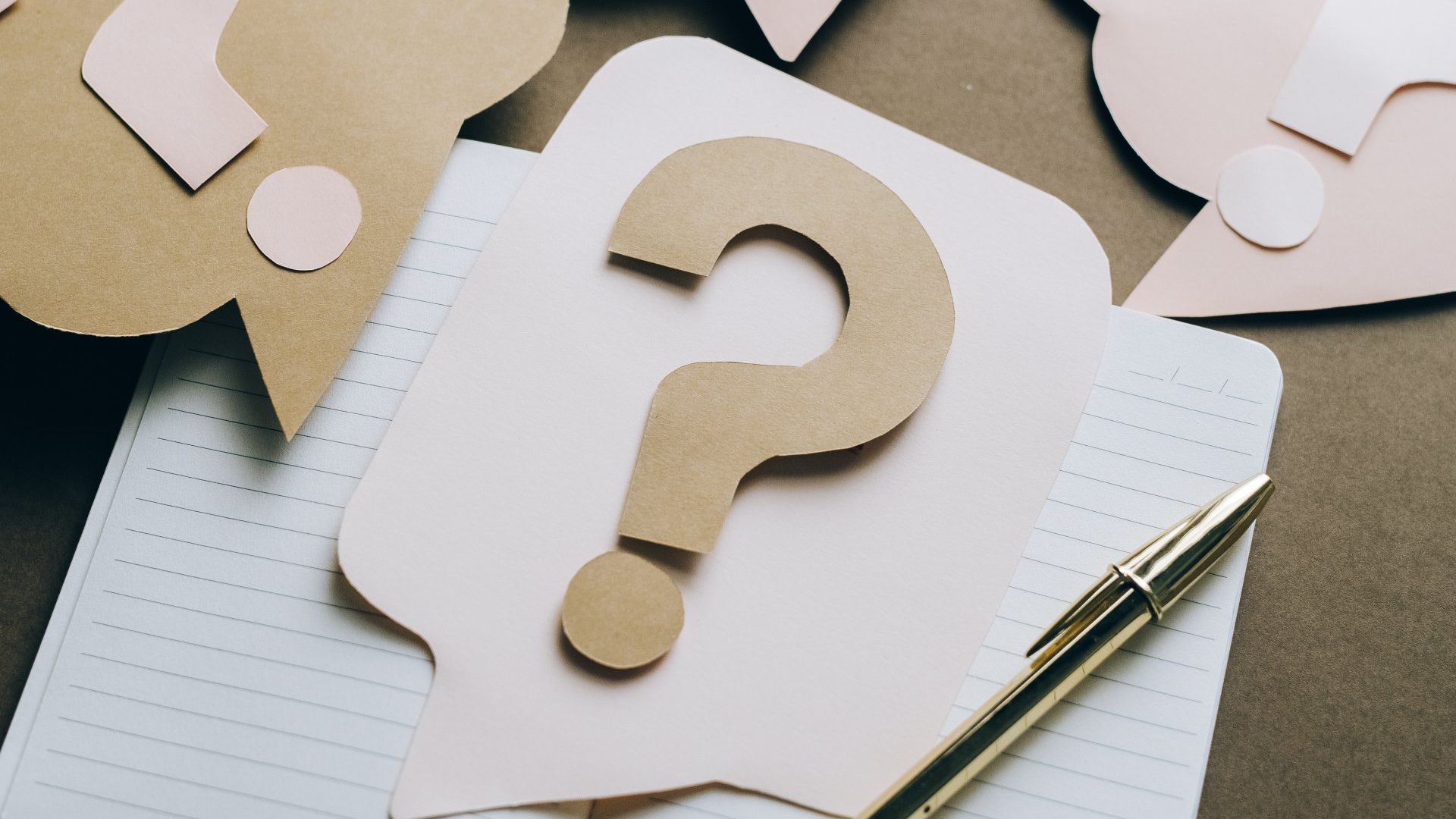 A paper question mark is sitting on top of a notebook next to a pen.