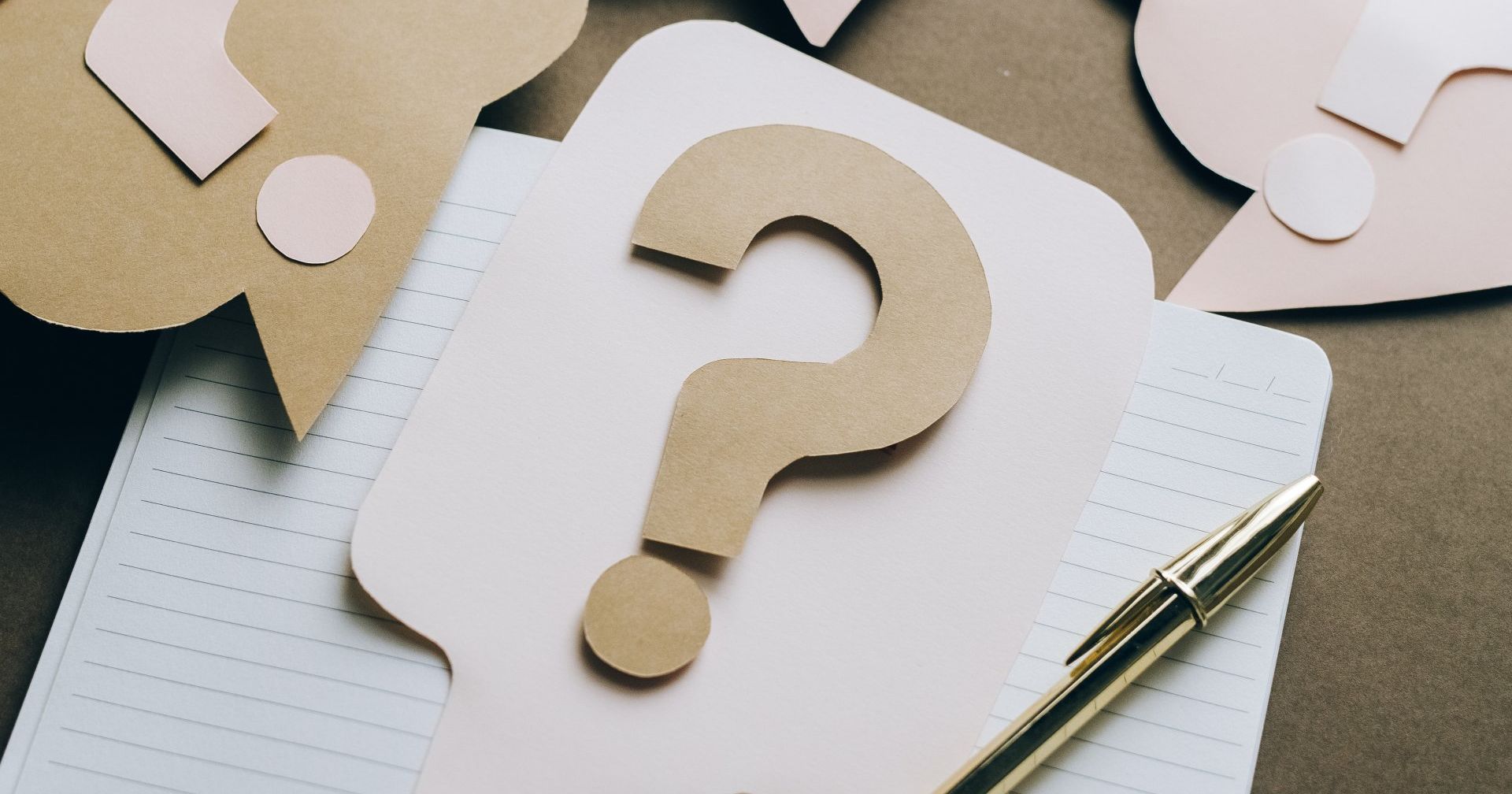A paper question mark is sitting on top of a notebook next to a pen.