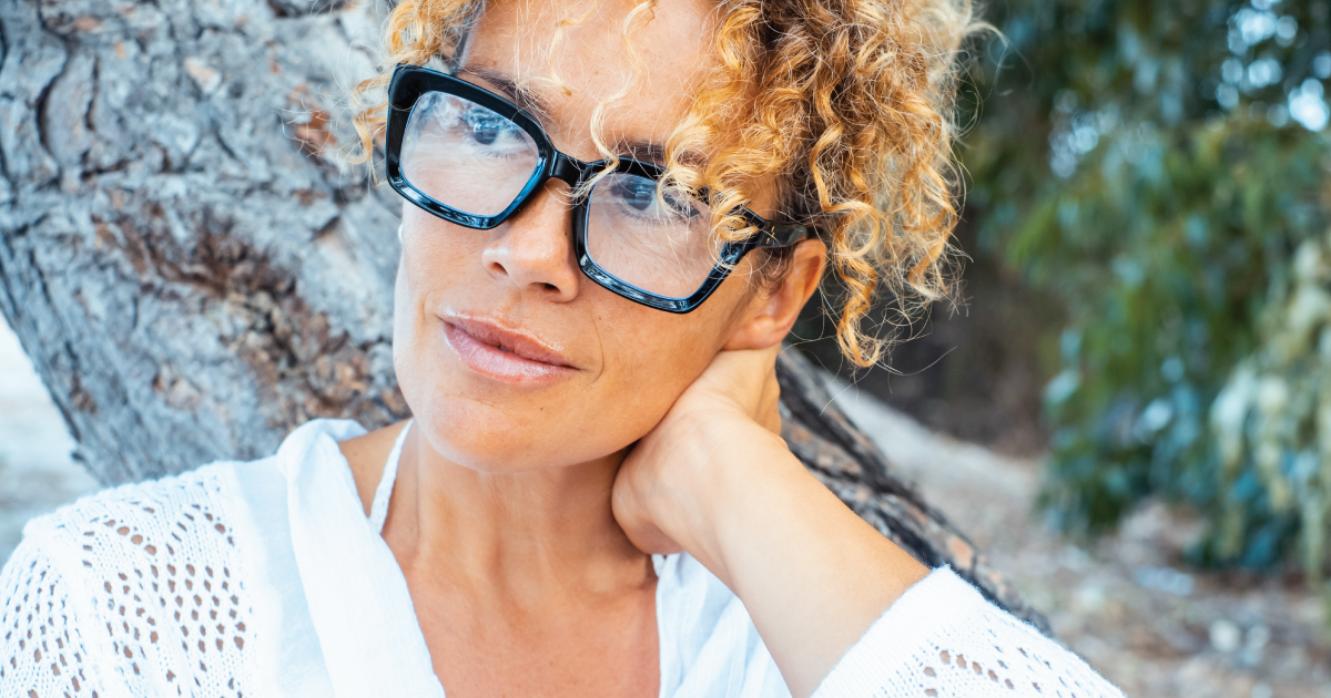 Close-up of a woman with curly hair and glasses, resting her hand on her neck outdoors