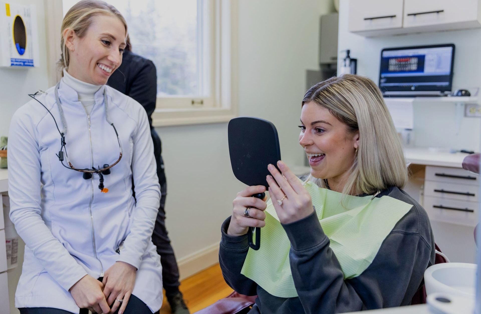 A woman is sitting in a dental chair looking at her teeth in a mirror.