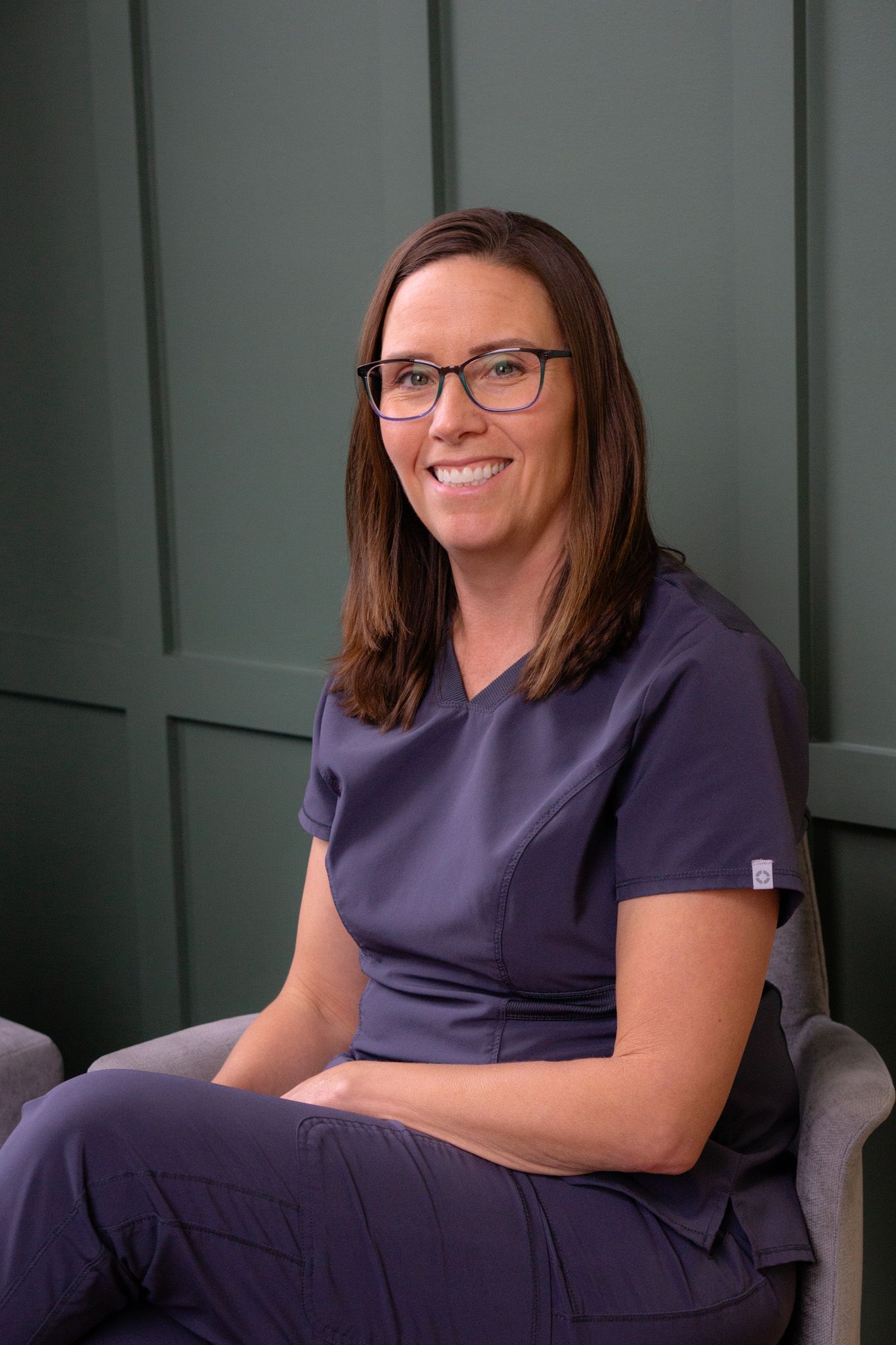 A woman wearing scrubs and glasses is sitting in a chair.