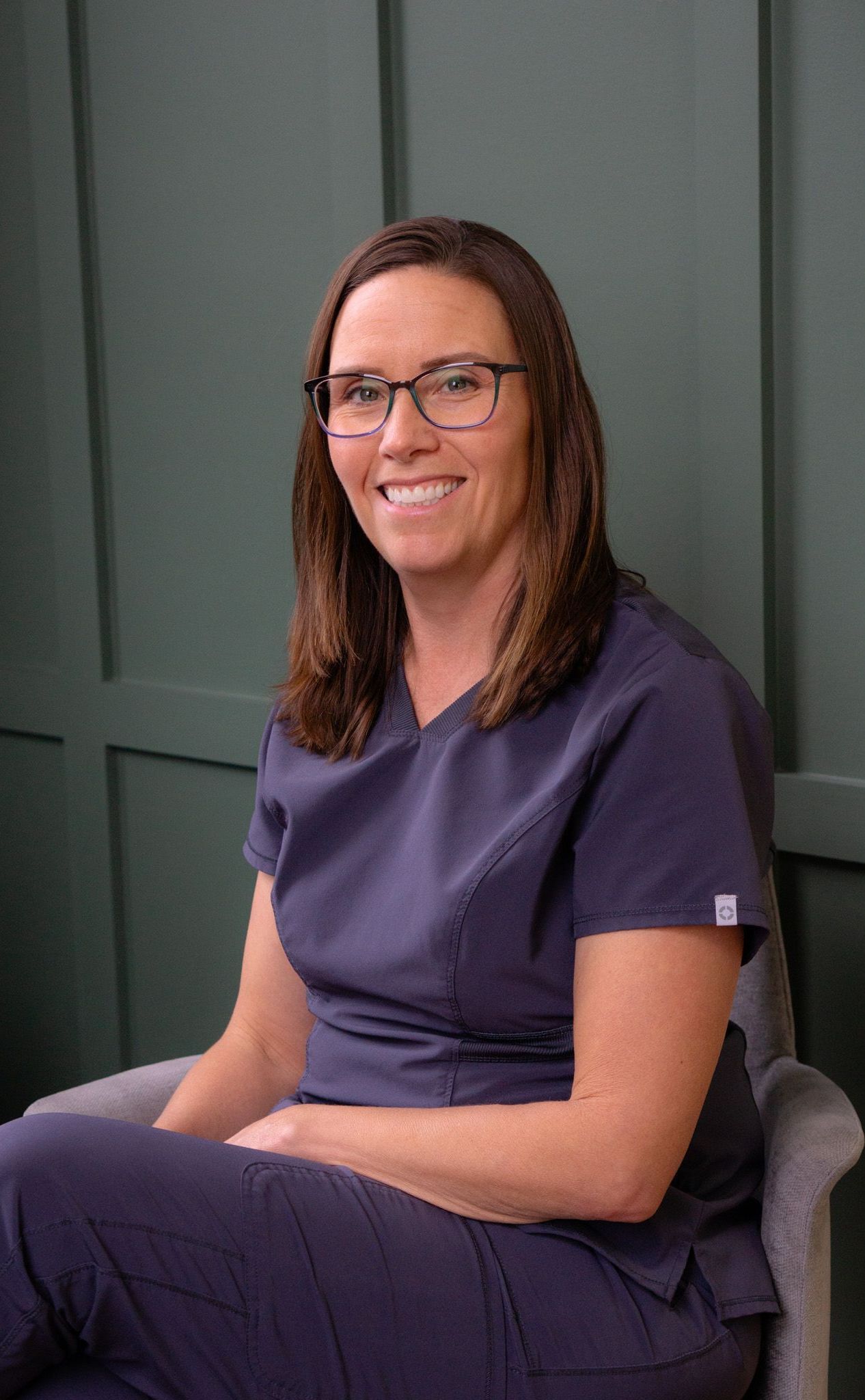 A woman wearing scrubs and glasses is sitting in a chair.