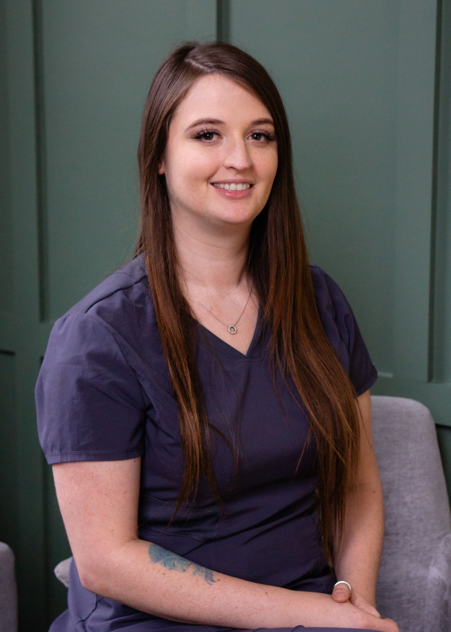 A woman in a purple scrub top is sitting in a chair and smiling.