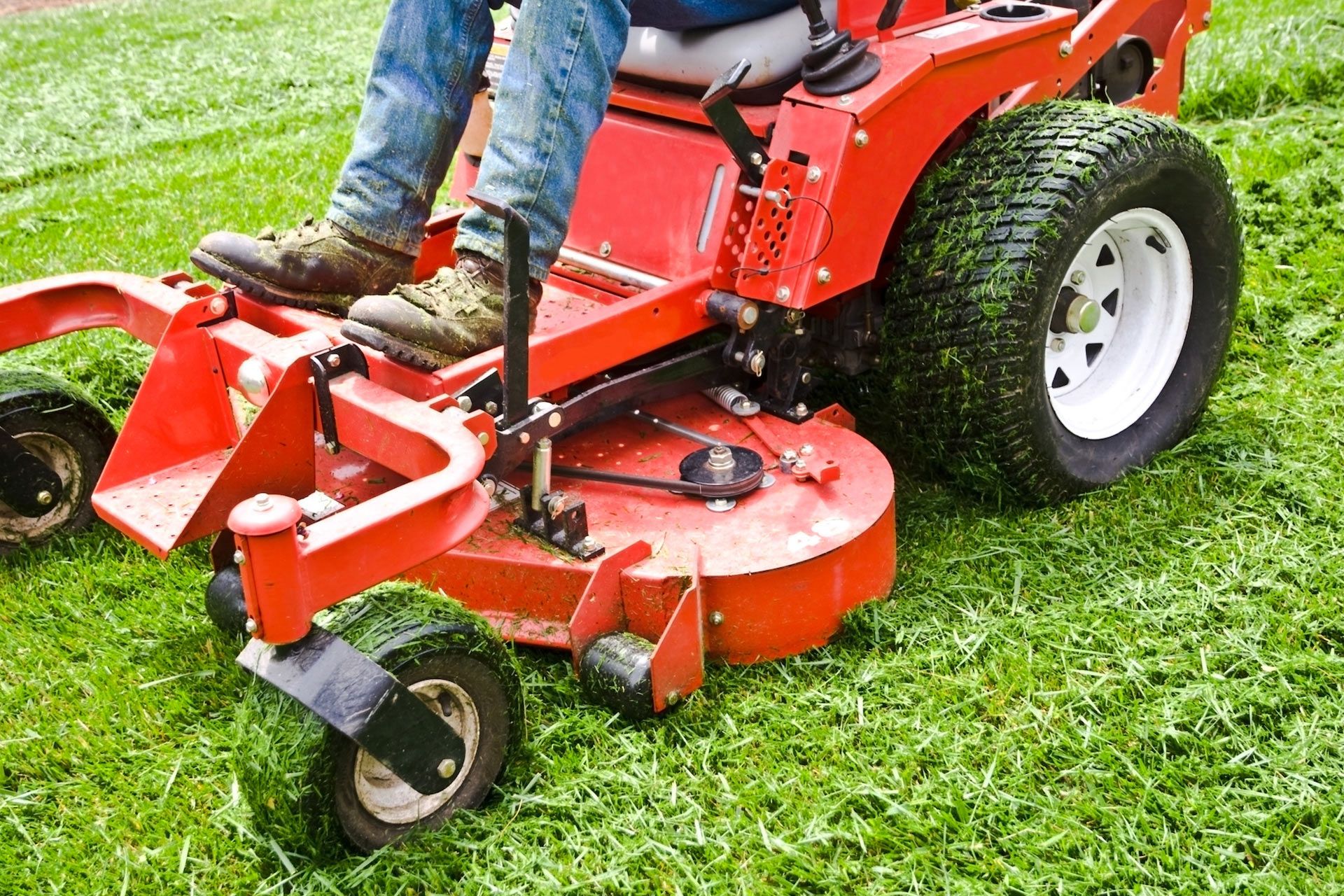 Person in blue jeans mowing green grass with a red zero-turn mower.