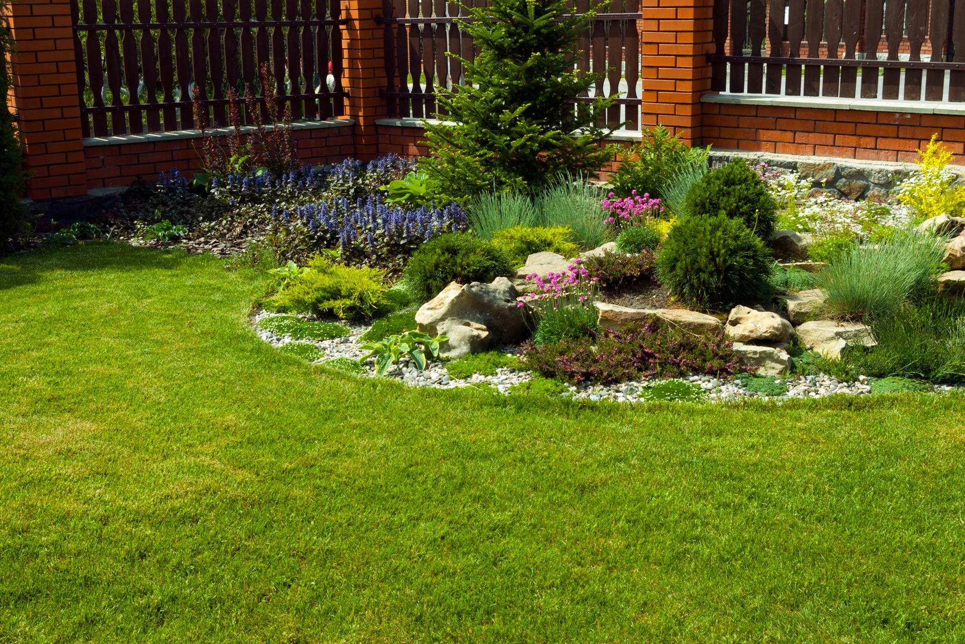 Lush green lawn borders a landscaped rock garden with various plants and a brick house.
