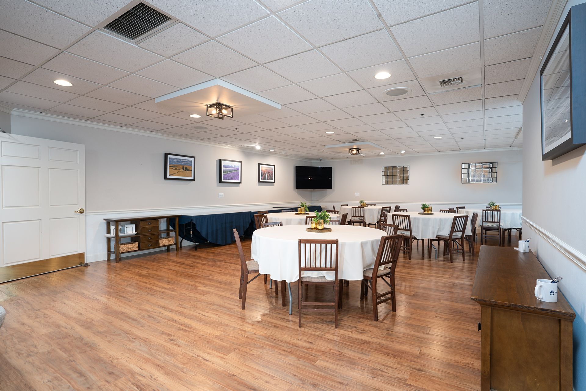 Bright dining room with round tables, wooden chairs, TV, framed wall art, and wood floors