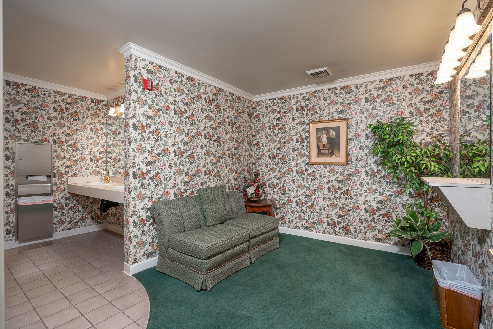 Small waiting room with floral wallpaper, green carpet, gray sofa, sink alcove, and potted plant.