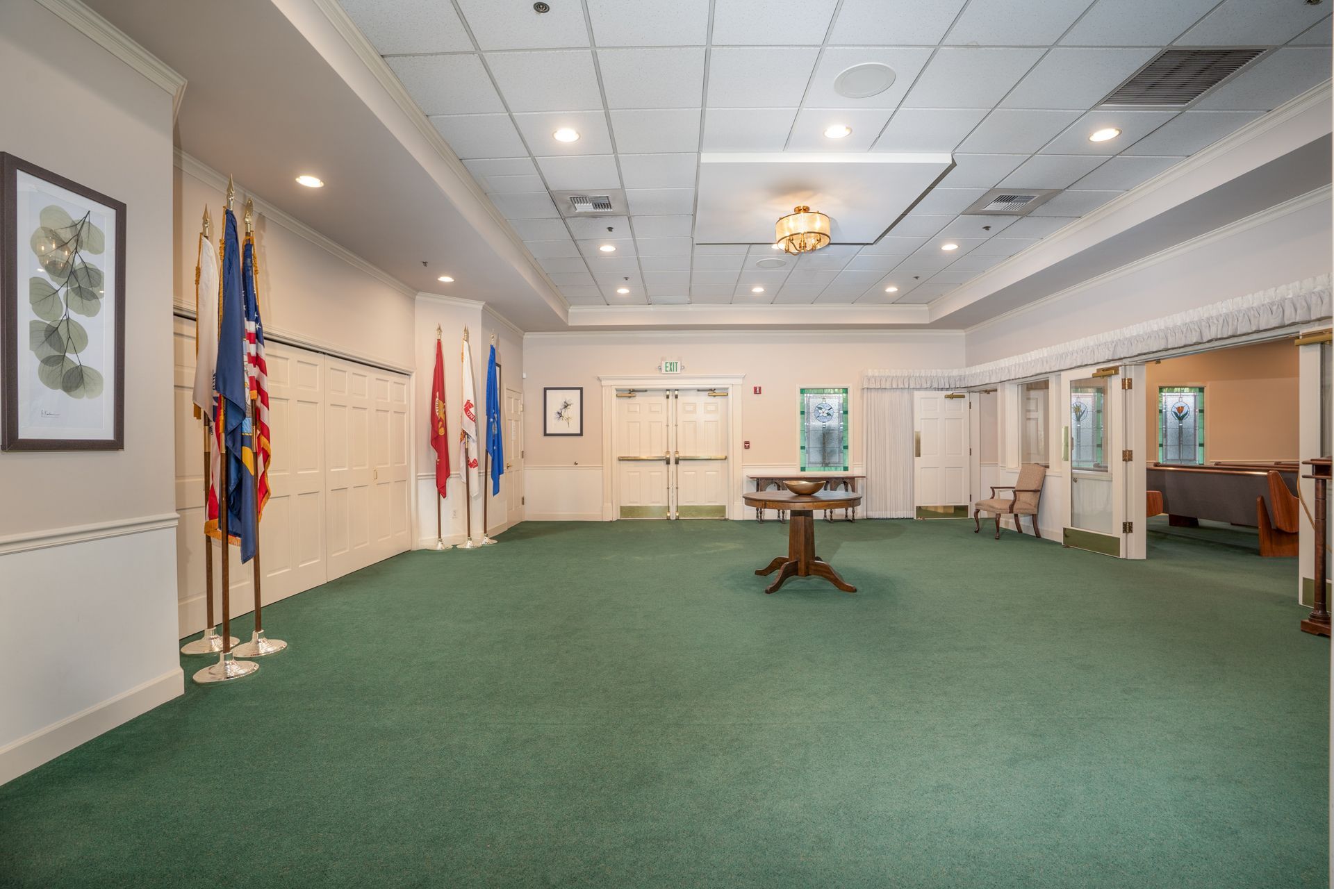 Empty carpeted meeting room with white walls, flags, and a central wooden table under recessed ceiling lights