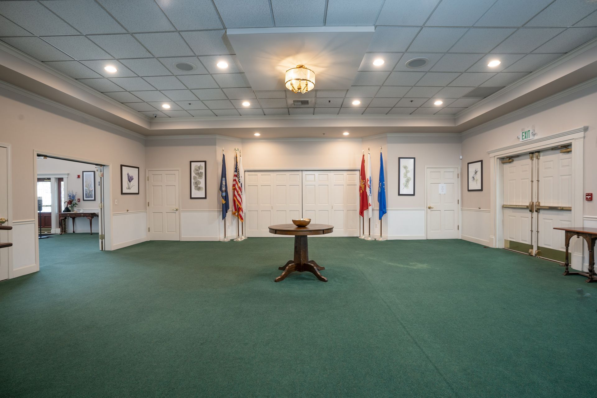 Empty conference room with green carpet, chandeliers, flags, and a small table centered beneath a whiteboard.