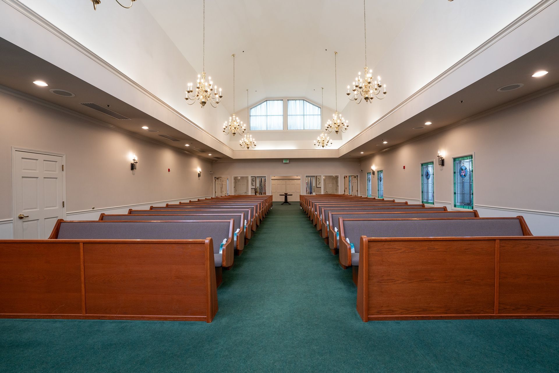 Empty church sanctuary with rows of wooden pews, green carpet, and chandeliers under a vaulted ceiling