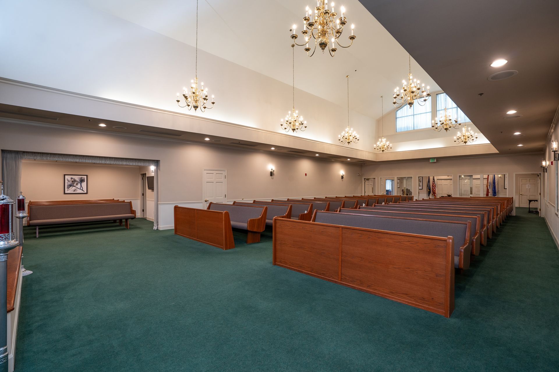 Empty church sanctuary with green carpet, wooden pews, chandeliers, and arched windows.