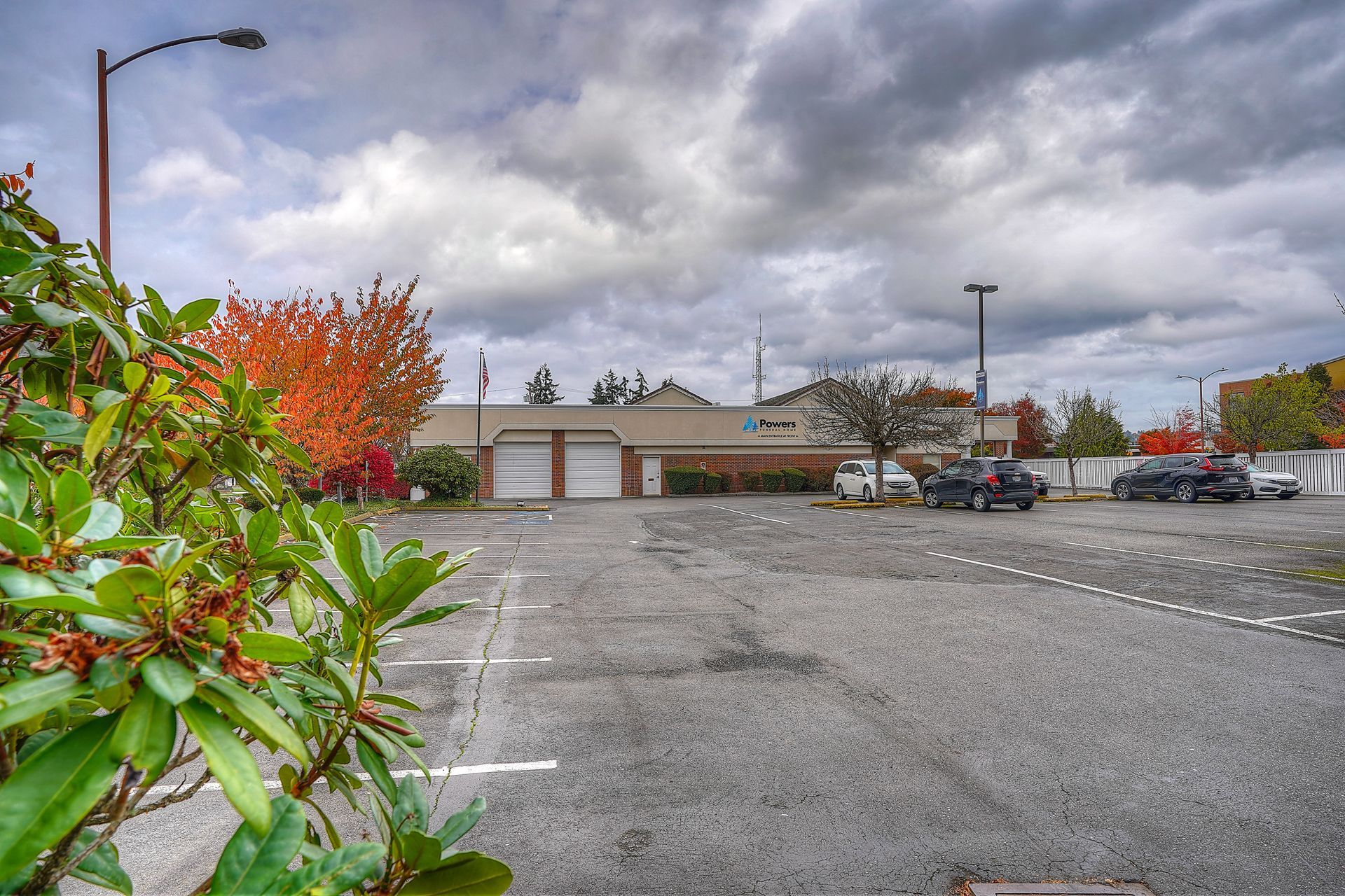 Empty parking lot with a low building, cloudy sky, and green shrubbery in the foreground