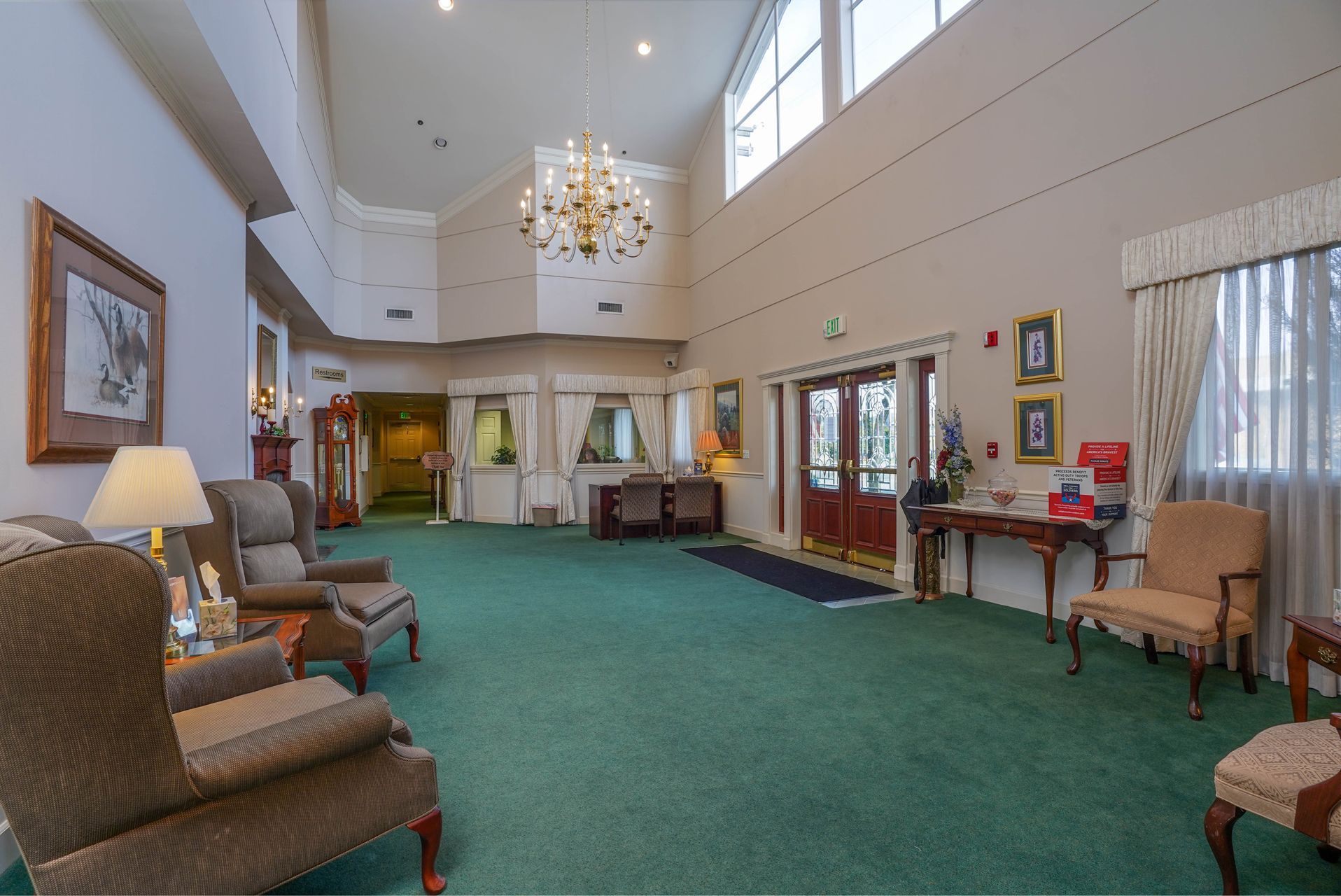 Spacious lounge with green carpet, high ceiling, chandelier, chairs, and large windows along the wall