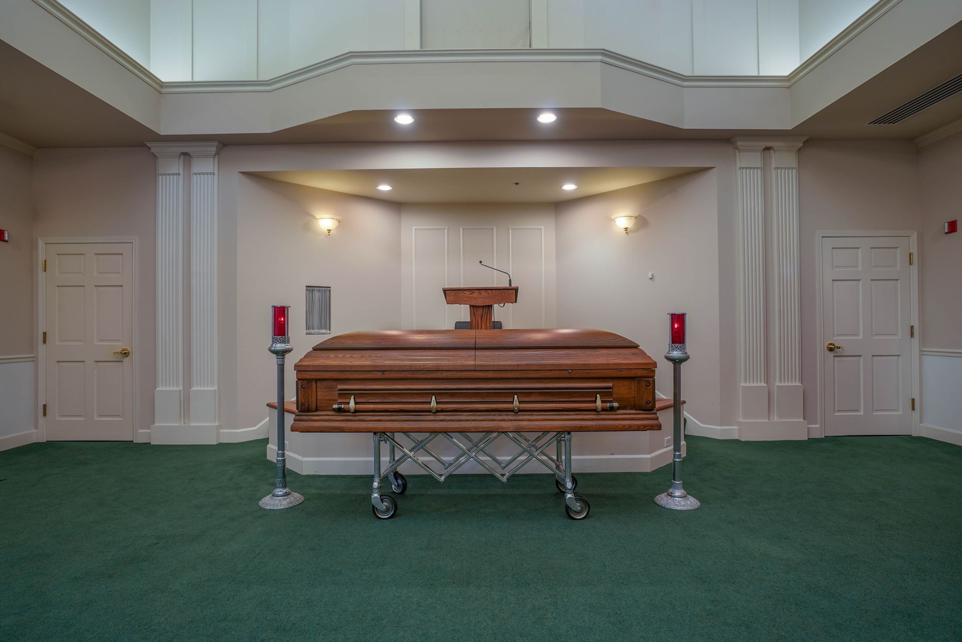 Wooden casket in a chapel with white walls and green carpet, lit by soft ceiling lights.