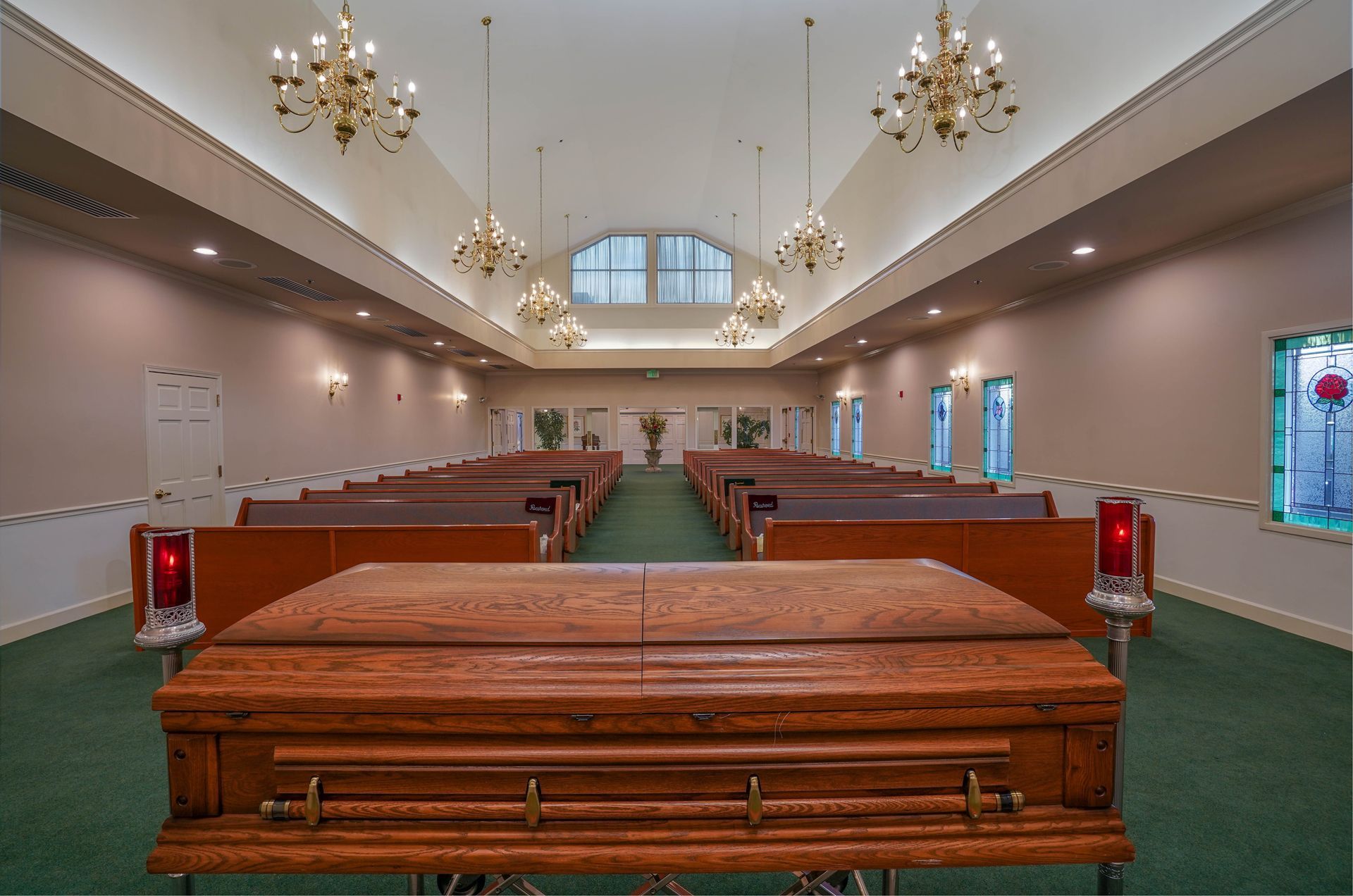 Empty funeral chapel with polished wooden caskets, green carpet, and chandeliers under a high arched ceiling