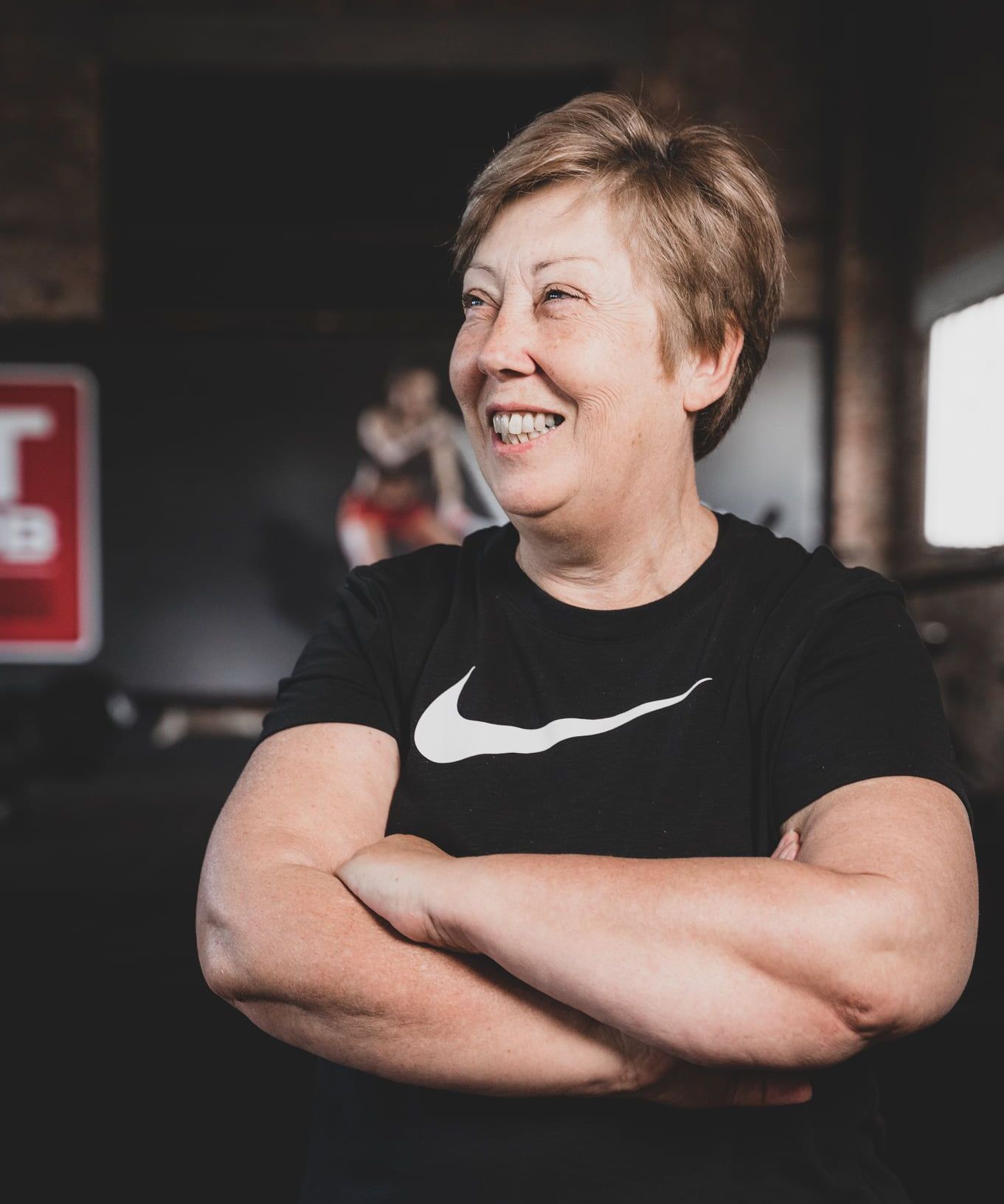 Een lachend persoon in een zwart Nike T-shirt met de armen over elkaar, staand in een ruimte die op een sportschool lijkt.