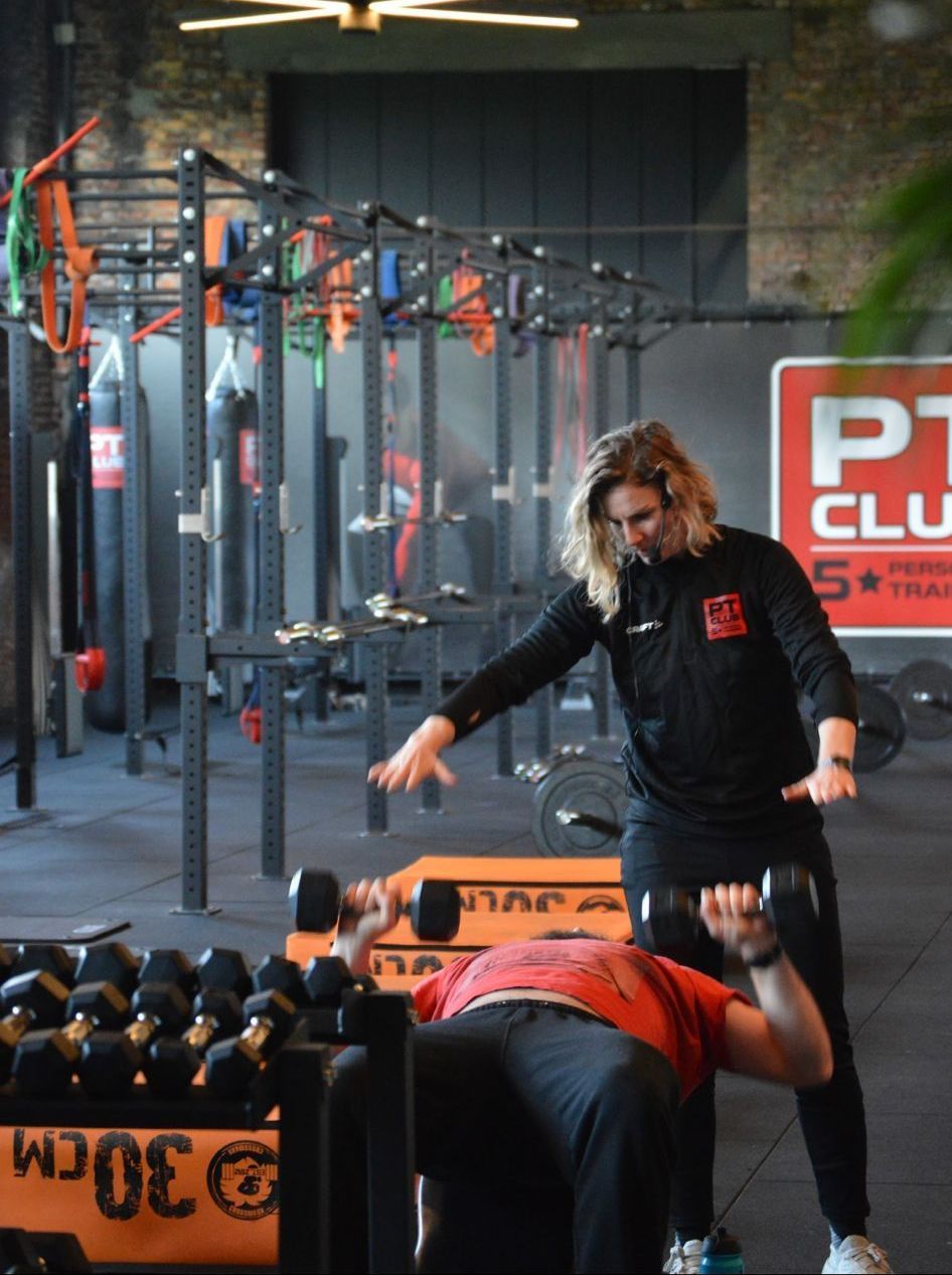 Een trainer begeleidt iemand bij het bankdrukken in een sportschool met dumbbells en squat racks.