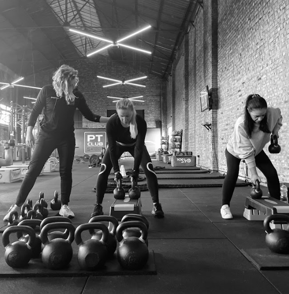 Drie mensen doen kettlebell-oefeningen in een sportschool met rijen gewichten voor zich. krachttraining hasselt