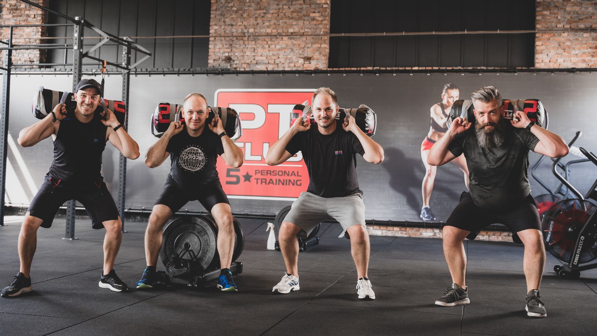 Vijf mensen doen een synchrone squat-workout in een sportschool met een rode banner op de achtergrond.