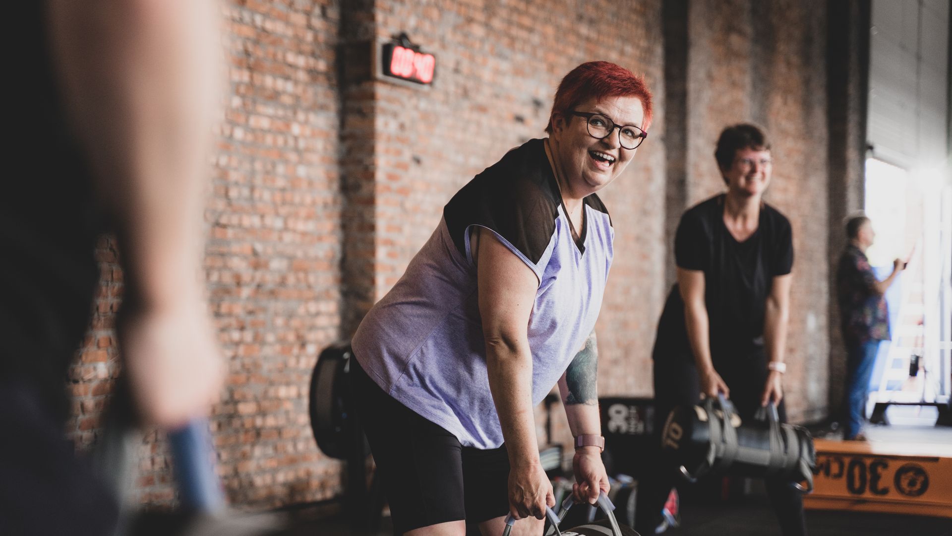 Een vrouw tilt gewichten in een sportschool, terwijl anderen op de achtergrond aan het sporten zijn.