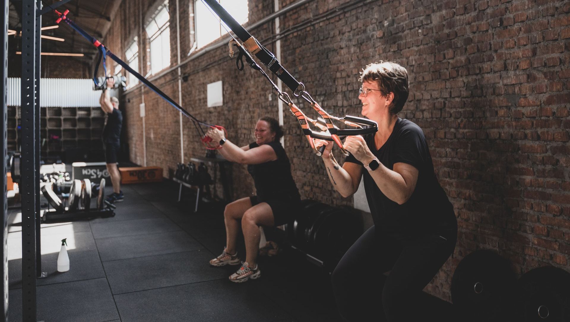 Twee mensen doen TRX-oefeningen in een sportschool met bakstenen muren.